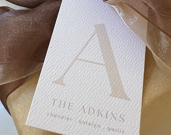 May include: A white gift tag with a textured surface. The tag features a large letter "A" in a light brown color. The text "THE ADKINS" is printed in a light brown color below the letter. The names "chandler", "katelyn", and "gotlin" are printed in a smaller font below the text.