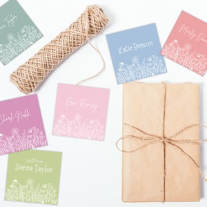 May include: Five gift tags with floral designs and names written on them. The tags are in various colors, including green, pink, blue, and light pink. The tags are arranged on a white surface with a roll of twine.