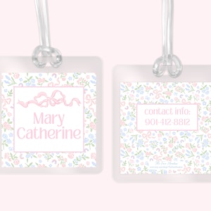 May include: A white plastic luggage tag with a floral pattern and pink accents. The tag has a clear plastic strap and a pink ribbon bow. The text "Mary Catherine" is printed in pink on the tag. The back of the tag has the text "contact info: 901-412-8812" printed in pink.