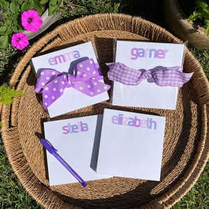 May include: Four personalized notepad sets with purple and white bows. The notepads are white with a purple ombre design and the names "Emma", "Garner", "Stella", and "Elizabeth" are printed on them.
