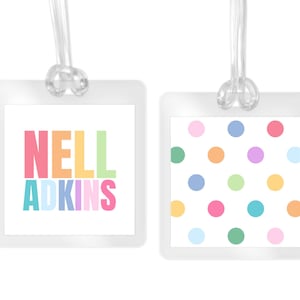 May include: Two clear luggage tags with a clear loop. One tag has the name "NELL ADKINS" in colorful letters. The other tag has a white background with colorful polka dots. Both tags are square.