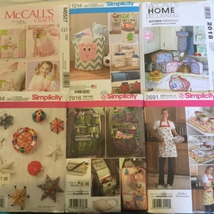 May include: A collection of Simplicity and McCall's sewing pattern envelopes. The patterns feature designs for home decor, kitchen essentials, and crafts. The envelopes display images of finished projects, including aprons, storage bags, and decorative items.