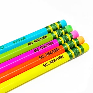 Puede incluir: Cinco lápices de colores con gomas rayadas verdes y amarillas. Los lápices están etiquetados con "MS. NGUYEN" en letras marrones. Los lápices están dispuestos en diagonal de arriba a la izquierda a abajo a la derecha.
