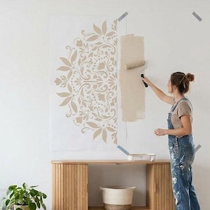 Könnte beinhalten: Ein helles Wohnzimmer mit grauem Ecksofa, Holztisch und dekorativer Wandvorlage. Eine Person bemalt einen Teil der Wand mit einer Rolle. Der Raum hat große Fenster und wirkt hell und luftig.