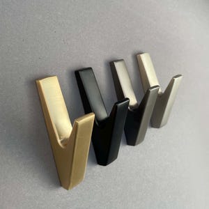 Op de afbeelding: Vier wandhaken in verschillende afwerkingen: goud, zwart, nikkel en geborsteld nikkel. De haken zijn V-vormig en zijn gemonteerd op een grijze muur.