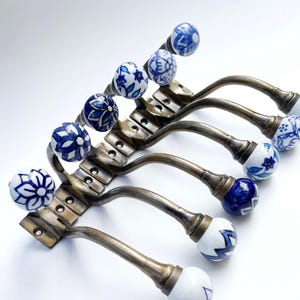 Può includere: Un set di sei ganci decorativi da parete. Ogni gancio presenta un braccio curvo in metallo color bronzo e un pomello in ceramica. I pomelli sono bianchi con motivi floreali blu. I ganci sono montati su una base metallica rettangolare.