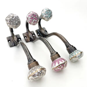 Könnte beinhalten: Vier dekorative Wandhaken mit verzierten Keramikknöpfen mit floralen Mustern in Grau-, Rosa- und Türkistönen. Die Haken haben eine Bronzefinish und sind auf quadratischen Metallsockeln montiert.