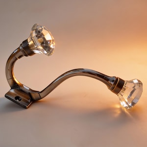 Könnte beinhalten: Ein Wandhaken aus Metall mit antikem Bronze-Finish, der zwei geschwungene Arme besitzt, die jeweils in einem facettierten, klaren Glaskristallknopf enden. Die Basis verfügt über ein Schraubloch zur Montage an einer flachen Oberfläche.