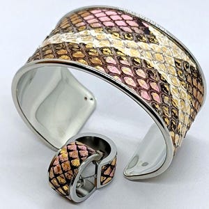May include: A stainless steel cuff bracelet and ring set. The bracelet and ring feature a snakeskin pattern in shades of pink, gold, and brown. The jewelry is set against a white background.