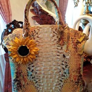 Puede incluir: Un bolso de mano de cuero multicolor con estampado de cocodrilo. El bolso tiene un asa superior y un dije de girasol desmontable. El bolso tiene un logotipo de metal dorado en la parte delantera.
