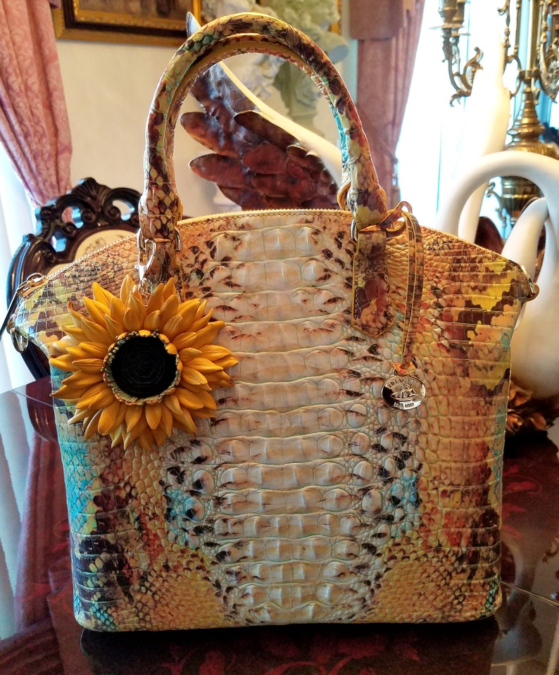 Puede incluir: Un bolso de mano de cuero multicolor con estampado de cocodrilo. El bolso tiene un asa superior y un dije de girasol desmontable. El bolso tiene un logotipo de metal dorado en la parte delantera.