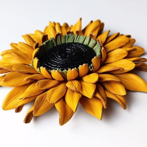 Puede incluir: Un broche de girasol de cuero amarillo con un centro verde y un detalle negro en el centro.