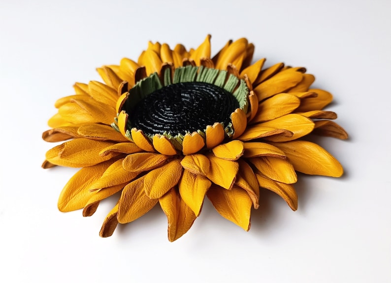Puede incluir: Un broche de girasol de cuero amarillo con un centro verde y un detalle negro en el centro.