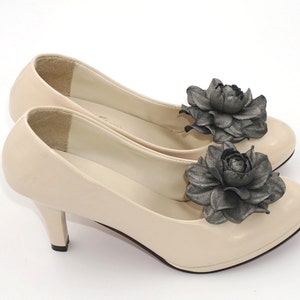 K&ouml;nnte beinhalten: Ein Paar beigefarbene Pumps mit hohen Abs&auml;tzen und einer schwarzen Blumenverzierung an jedem Schuh.