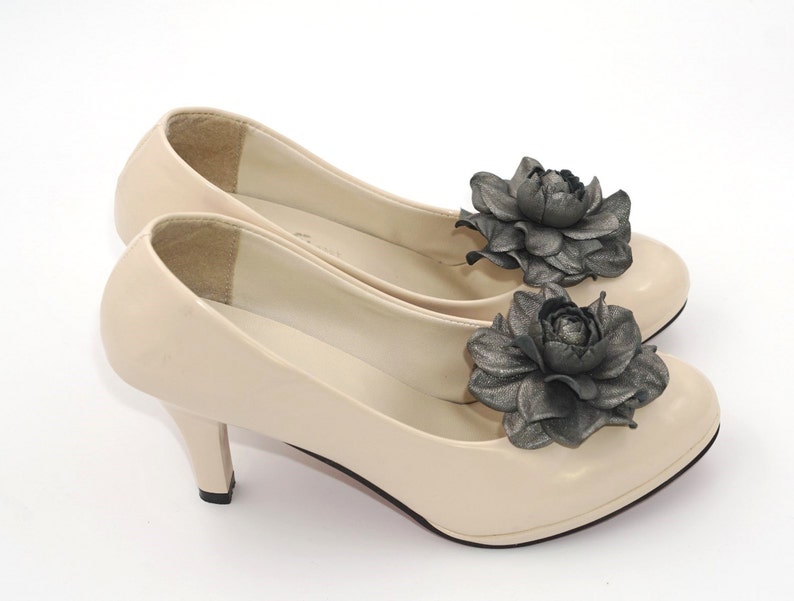 K&ouml;nnte beinhalten: Ein Paar beigefarbene Pumps mit hohen Abs&auml;tzen und einer schwarzen Blumenverzierung an jedem Schuh.