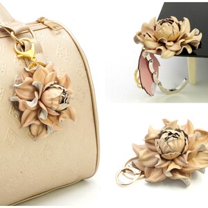 May include: A beige leather flower keychain with gold-colored hardware. The flower has multiple layers of petals and is attached to a gold-colored clasp. The keychain is shown attached to a beige handbag.