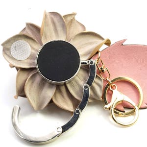 May include: A silver and black purse hook with a gold-colored keychain and clasp. The hook has a circular black top and a folding design. It is displayed on a decorative flower and a pink leaf.