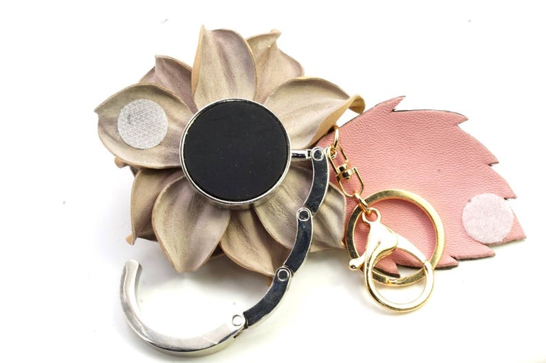 May include: A silver and black purse hook with a gold-colored keychain and clasp. The hook has a circular black top and a folding design. It is displayed on a decorative flower and a pink leaf.