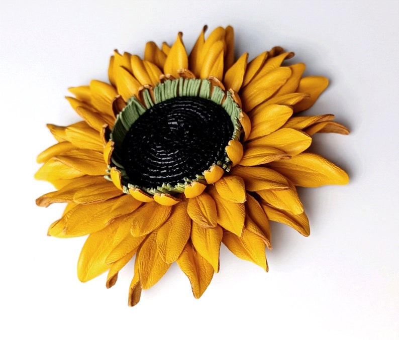Puede incluir: Un broche de girasol de cuero amarillo con un centro negro y detalles verdes. El girasol est&aacute; hecho de p&eacute;talos de cuero individuales.