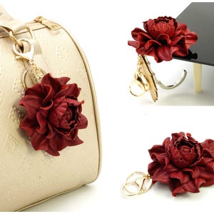 May include: A burgundy leather flower keychain with gold-colored hardware. The flower is a detailed peony shape, and the keychain is attached to a gold-colored purse. The background is white.