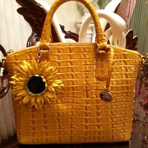 Puede incluir: Un bolso de mano de cuero amarillo con un patr&oacute;n de cocodrilo en relieve y un dije de girasol. El bolso tiene dos asas superiores y un cierre de cremallera.