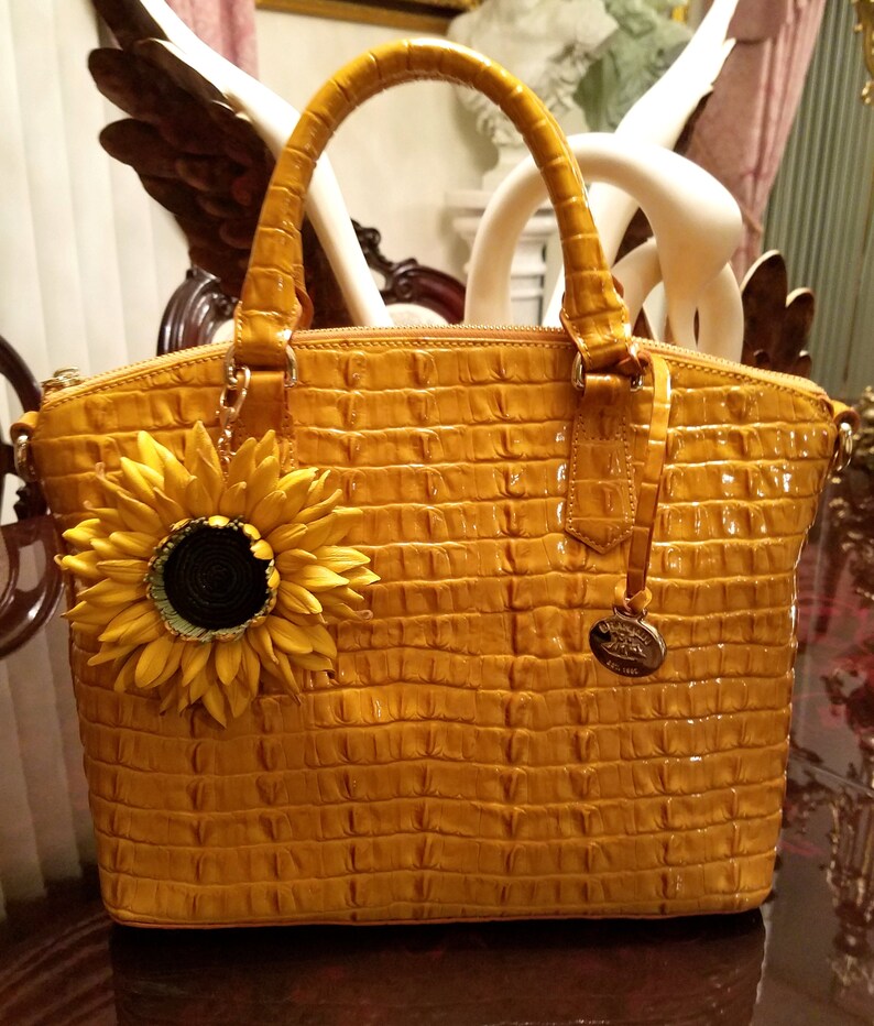 Puede incluir: Un bolso de mano de cuero amarillo con un patr&oacute;n de cocodrilo en relieve y un dije de girasol. El bolso tiene dos asas superiores y un cierre de cremallera.