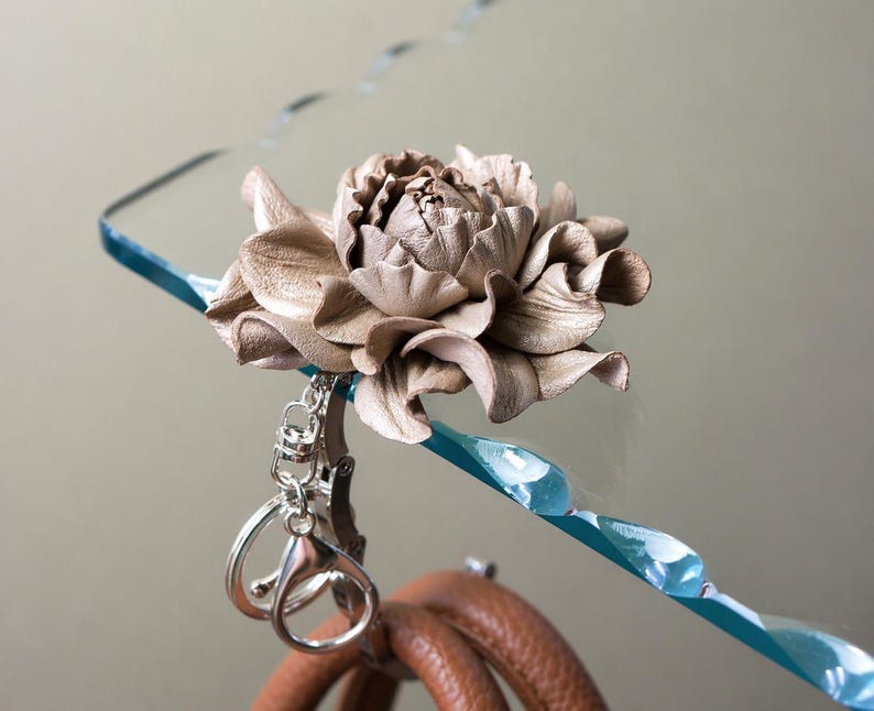 May include: A handcrafted leather flower in a neutral tone, attached to a silver keychain. The flower is intricately detailed, with layered petals. A blue, twisted accent piece is visible in the background, along with a brown leather item.