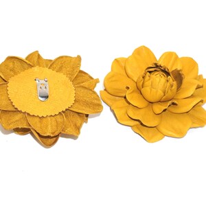 May include: Two yellow leather flower brooches. One brooch is shown from the back, revealing a metal clip. The other brooch is shown from the front.