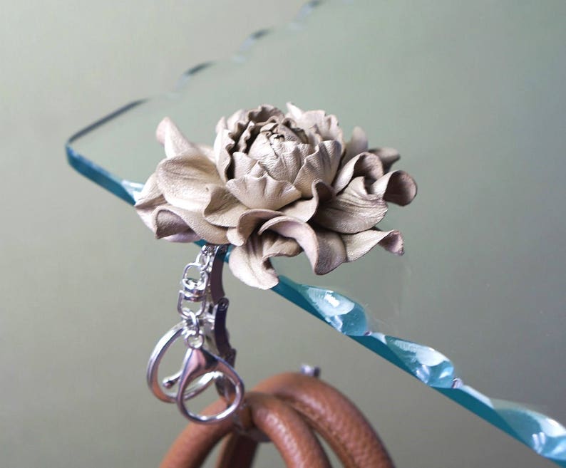 May include: A handcrafted, light beige leather flower key chain. The flower is attached to a silver-colored metal clasp, with a brown leather handle visible below. The flower is detailed with realistic petal textures.