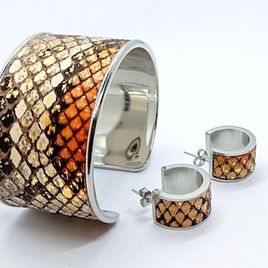 Puede incluir: Un brazalete ancho y pendientes de aro a juego. La joyería presenta un patrón de piel de serpiente en tonos marrón, naranja y dorado, sobre metal plateado. Los pendientes son aros pequeños.