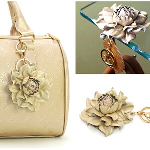 May include: A beige leather flower keychain with a gold-colored key ring and clasp. The flower is attached to a gold-colored feather charm. The keychain is shown attached to a beige handbag.