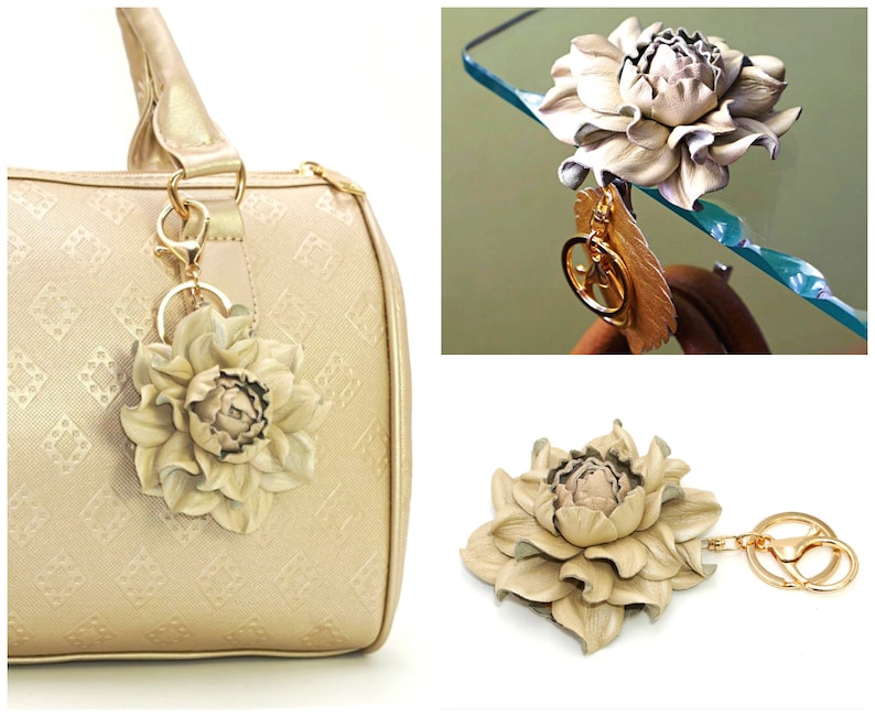 May include: A beige leather flower keychain with a gold-colored key ring and clasp. The flower is attached to a gold-colored feather charm. The keychain is shown attached to a beige handbag.