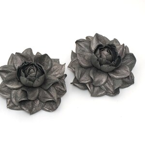 K&ouml;nnte beinhalten: Zwei graue Lederblumenbroschen mit einem 3D-Design. Die Blumen haben ein mehrschichtiges Bl&uuml;tenblattdesign und eine zentrale Knospe.