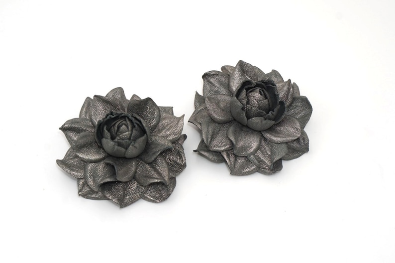 K&ouml;nnte beinhalten: Zwei graue Lederblumenbroschen mit einem 3D-Design. Die Blumen haben ein mehrschichtiges Bl&uuml;tenblattdesign und eine zentrale Knospe.
