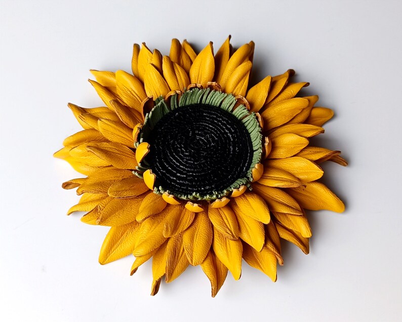 Puede incluir: Un broche de girasol de cuero amarillo con un centro verde. El girasol est&aacute; hecho de p&eacute;talos individuales cosidos juntos.