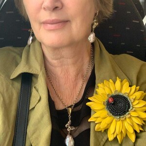Puede incluir: Una mujer lleva una chaqueta verde con un broche de girasol, un collar de perlas con un colgante de mariposa y pendientes de perlas con detalles de mariposa.