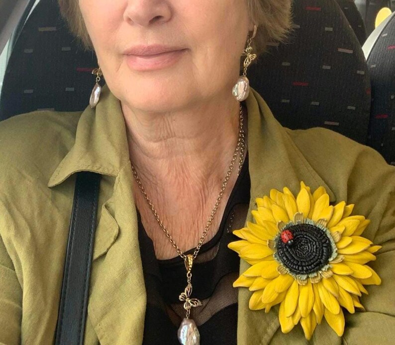 Puede incluir: Una mujer lleva una chaqueta verde con un broche de girasol, un collar de perlas con un colgante de mariposa y pendientes de perlas con detalles de mariposa.