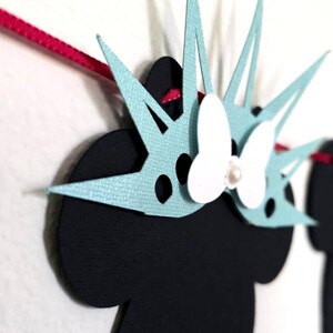 May include: A decorative banner featuring black silhouettes with light blue and white accents. The banner includes a Minnie Mouse-inspired design with a bow and pearl, and other patriotic-themed shapes. The banner is strung on a pink ribbon.