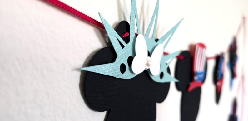 May include: A decorative banner featuring black silhouettes with light blue and white accents. The banner includes a Minnie Mouse-inspired design with a bow and pearl, and other patriotic-themed shapes. The banner is strung on a pink ribbon.