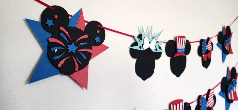 May include: A festive banner featuring Mickey Mouse silhouettes and star shapes in red, white, and blue. The banner includes designs of fireworks, the Statue of Liberty, and Uncle Sam hats, perfect for patriotic celebrations.