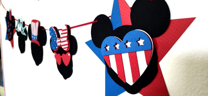 May include: A patriotic garland featuring Mickey Mouse-shaped cutouts in red, white, and blue. The garland includes stars, hearts, and bowties, perfect for Independence Day or other festive occasions. The design incorporates the colors of the American flag.