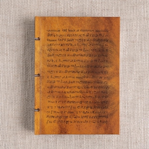 May include: A tan-colored journal with the text "THE BOOK OF MORMON" and other script. The cover is a warm, textured tan color, and the pages are bound with three metal rings. The text is written in black ink.