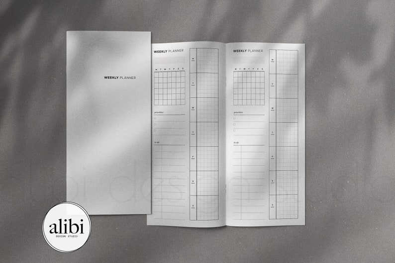 May include: A white weekly planner with a black grid and lines. The planner has two pages, each with a weekly layout. The planner is closed and has a white cover with the text "WEEKLY PLANNER" printed in black.