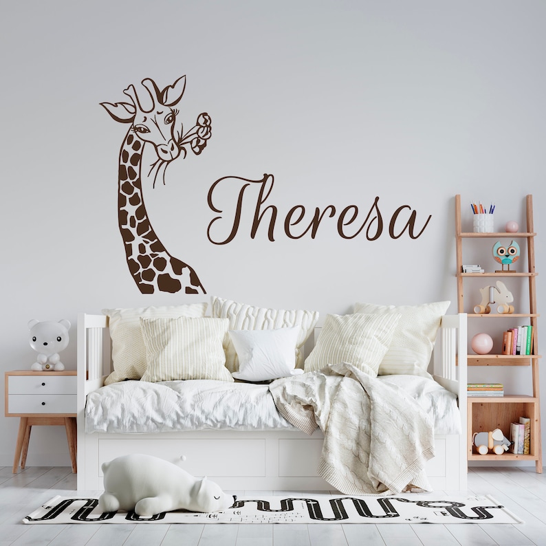 May include: A brown giraffe silhouette wall decal with flowers in its mouth, above a white bed with white bedding and pillows. The name "Theresa" is written in cursive script below the giraffe.