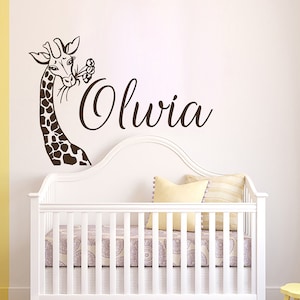 May include: A brown giraffe wall decal with a flower in its mouth, above a white crib with a white and gray patterned sheet. The name "Olivia" is written in cursive script below the giraffe.