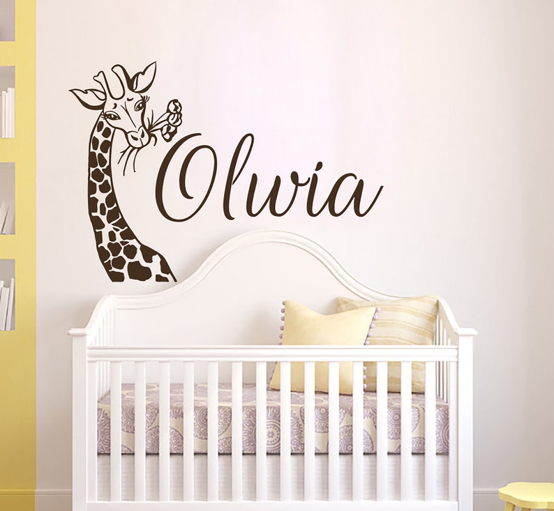 May include: A brown giraffe wall decal with a flower in its mouth, above a white crib with a white and gray patterned sheet. The name "Olivia" is written in cursive script below the giraffe.
