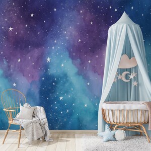 Pode incluir: Um quarto de criança com um mural de parede com estampa de galáxia em aquarela em tons de azul profundo e roxo com estrelas brancas. Um dossel de tecido azul claro pende sobre um berço de madeira com uma almofada de estrela e um cobertor.