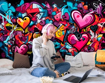 Puede incluir: Un colorido mural de graffiti con corazones y formas abstractas como telón de fondo. Una persona con una sudadera gris y vaqueros está sentada en una cama, con auriculares y sosteniendo un mando de videojuegos. También se ve un portátil y palomitas de maíz.