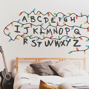 May include: Wall decal featuring the alphabet with string lights. The letters are in a dark gray font, and the string lights are in various colors, including red, green, blue, and yellow. The decal is applied to a white wall.