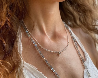 May include: A silver chain necklace with a small, iridescent gemstone pendant. The necklace is layered over a longer necklace made of iridescent beads.